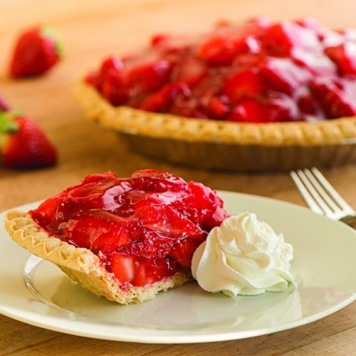 A slice of strawberry pie topped with glossy red glaze and whipped cream, served on a plate with a whole pie in the background.