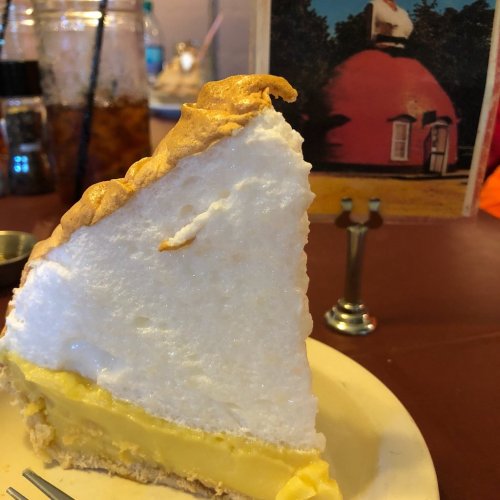 A slice of lemon meringue pie on a plate, with a vintage postcard in the background featuring a pink dome building.