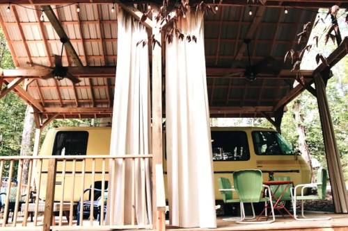 A cozy outdoor space with a yellow van, wooden deck, and curtains, surrounded by trees.