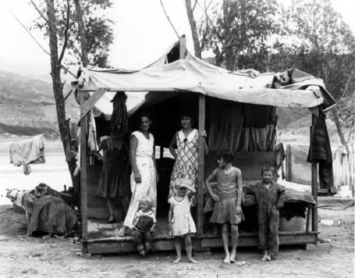 A black-and-white photo of a family outside a makeshift wooden shelter, with children and adults standing together.