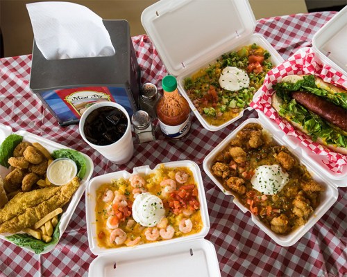 A spread of takeout food including shrimp, fried chicken, and a sausage sandwich on a checkered tablecloth.