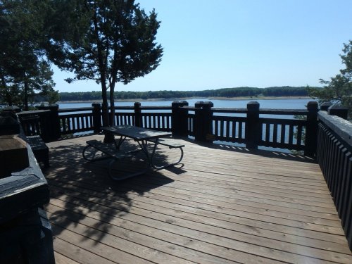 A wooden deck with a picnic table overlooks a calm lake surrounded by trees under a clear blue sky.
