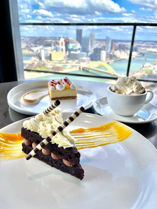 A slice of chocolate cake with cream and decorative stripes, alongside a dessert and a cup of whipped cream, with a city view.