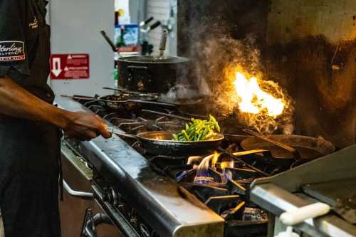 A chef stirs a pan on a busy stove, with flames and steam rising from the cooking surface. Green beans are visible in the pan.