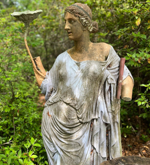A weathered statue of a woman holding a dish, draped in a flowing robe, surrounded by greenery.