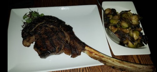 A large, grilled tomahawk steak on a plate, accompanied by a side of roasted Brussels sprouts.