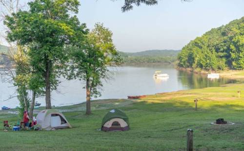 A serene lakeside campsite with tents, trees, and a boat on the water in the background.