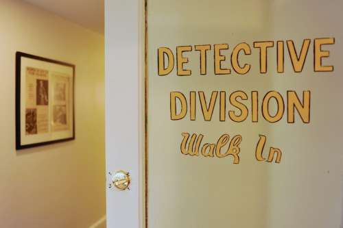A door with "Detective Division" and "Walk In" signs, leading to a hallway with framed artwork on the wall.