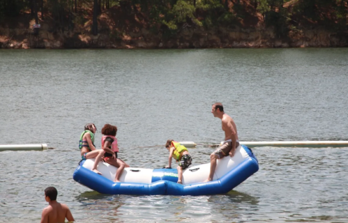 A group of children and an adult play on a floating inflatable in a lake on a sunny day.