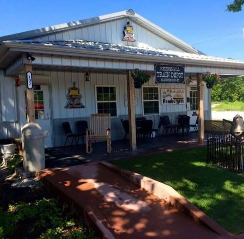 A quaint train depot building with a sign for Whisler's Hamburgers, featuring outdoor seating and flower pots.