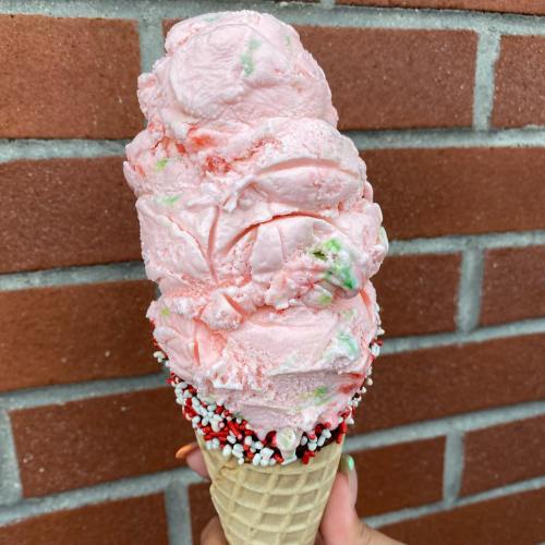 A large ice cream cone with pink mint-flavored scoops, topped with colorful sprinkles, against a brick wall.
