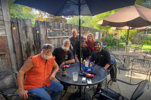 A group of six people sitting at a table outdoors, enjoying drinks under umbrellas in a casual setting.