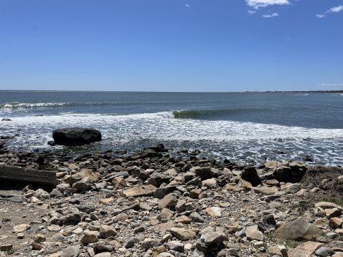 A rocky shoreline with gentle waves lapping at the water under a clear blue sky.