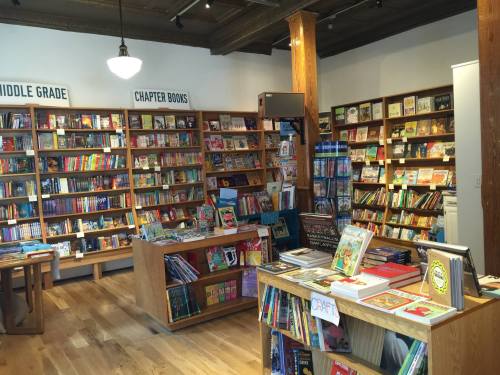 A cozy bookstore interior with wooden shelves filled with colorful books, including sections for middle grade and chapter books.