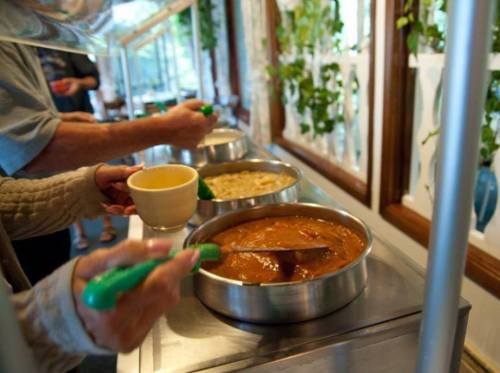 People serving food from a buffet, with bowls of soup and a thick sauce in metal containers. Green serving utensils are used.