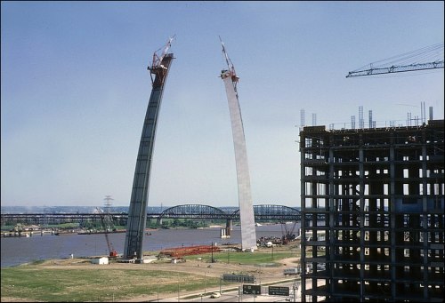 Construction of a large arch bridge over a river, with cranes and a building site in the foreground.