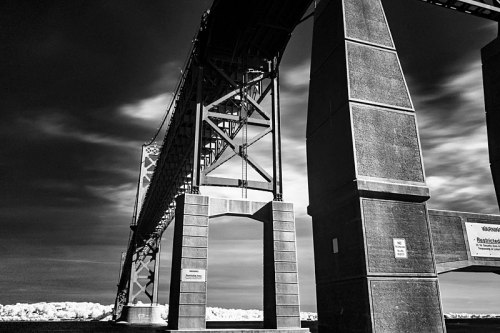 Black and white image of a large bridge structure with towering supports and dramatic cloud patterns in the sky.
