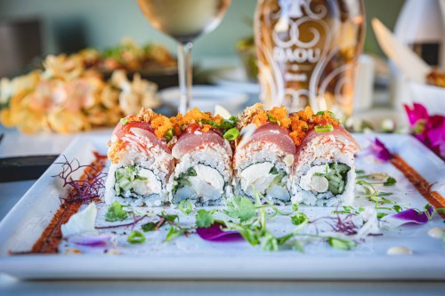 A close-up of sushi rolls topped with fish, roe, and garnishes, served on a white plate with flowers and a drink in the background.