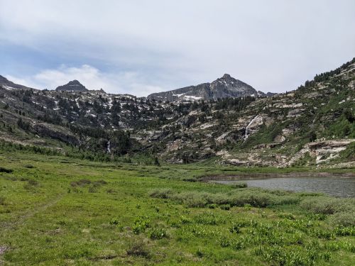 A scenic mountain landscape with green meadows, rocky peaks, and a calm lake under a partly cloudy sky.