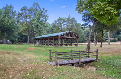 A grassy park with a wooden pavilion and a small bridge over a shallow area, surrounded by trees.