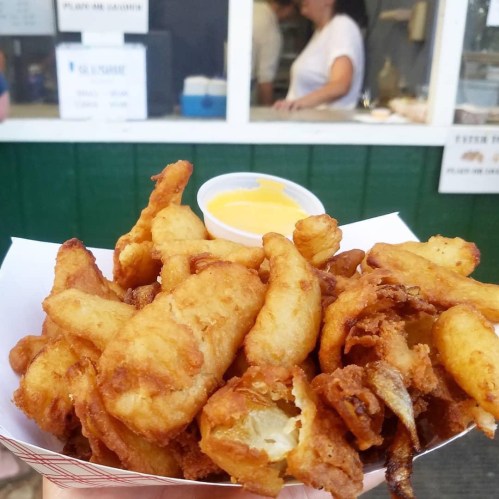 A serving of golden fried food with a side of dipping sauce, set against a blurred background of a food stand.