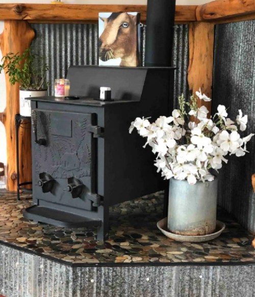 A black wood stove beside a metal bucket of white flowers, set against a rustic wooden and corrugated backdrop.
