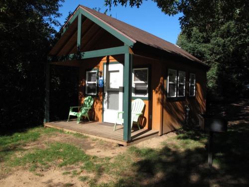 A small wooden cabin with a porch, featuring two green chairs and surrounded by trees under a clear blue sky.