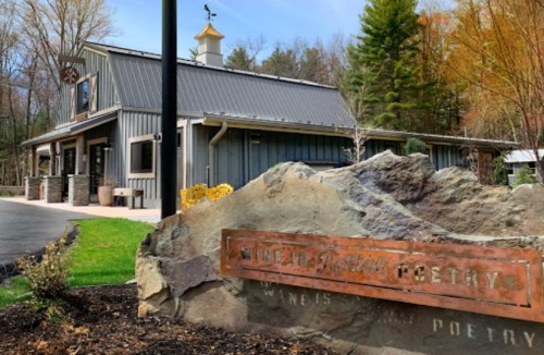 A rustic building with a metal roof, surrounded by trees, featuring a sign that reads "Wines & Poetry."