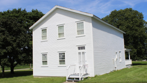 A white, two-story house with multiple windows and a front porch, set against a clear blue sky and green trees.