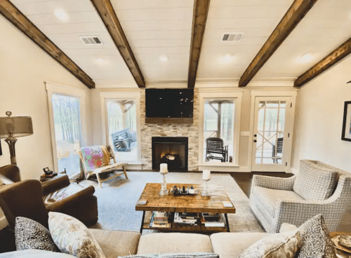 Cozy living room with a fireplace, wooden beams, and large windows overlooking a forested area.
