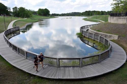 A curved wooden walkway along a calm lake, with two people jogging and reflections of clouds in the water.