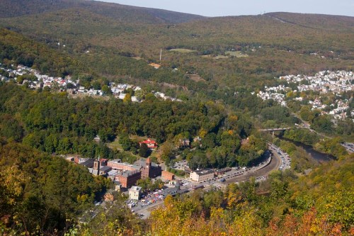 Aerial view of a valley with a town, surrounded by green hills and trees, with a winding road and river below.