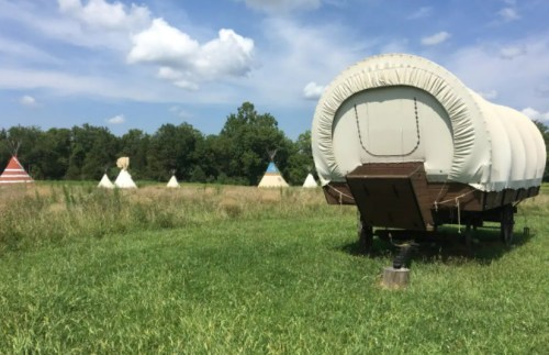 A covered wagon in a grassy field with several teepees in the background under a blue sky with clouds.
