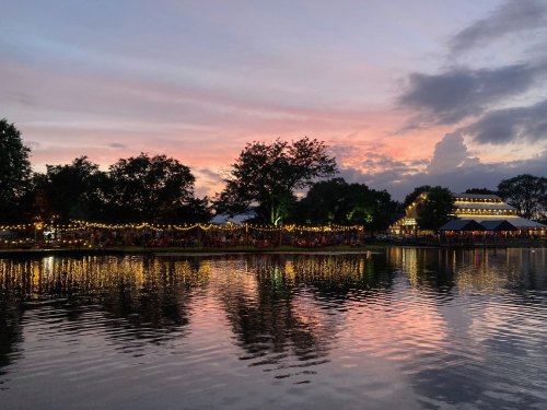 A serene lake at sunset, reflecting colorful skies and lights from nearby trees and buildings.