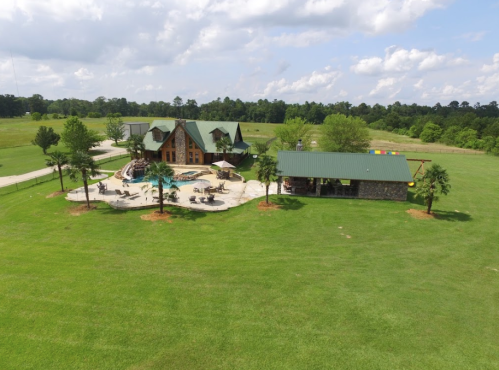 Aerial view of a large rustic house with a pool, surrounded by green grass and trees under a partly cloudy sky.