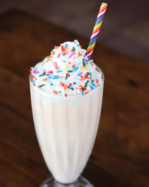 A tall glass of milkshake topped with whipped cream and colorful sprinkles, with a rainbow-striped straw.