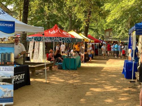 A vibrant outdoor market scene with colorful tents, vendors, and visitors enjoying the event under shaded trees.