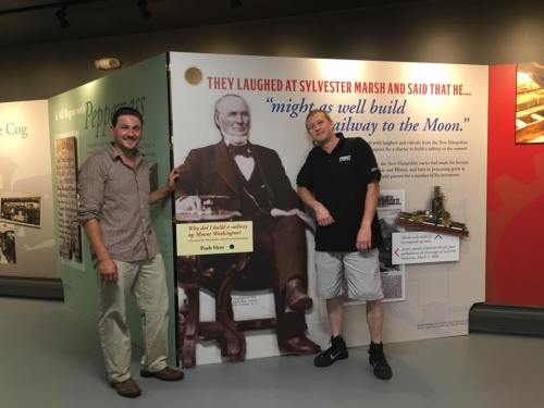 Two men pose next to a historical exhibit featuring a portrait of Sylvester Marsh and a quote about his ambitions.