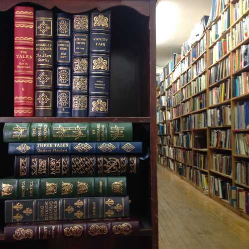 A wooden bookshelf filled with classic books, with a library aisle visible in the background.