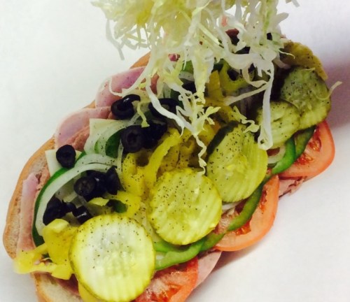A sub sandwich loaded with ham, pickles, olives, onions, green peppers, and shredded lettuce on a hoagie roll.