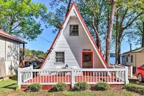 A charming A-frame cabin with a red roof, surrounded by trees and a small porch, near a lake.