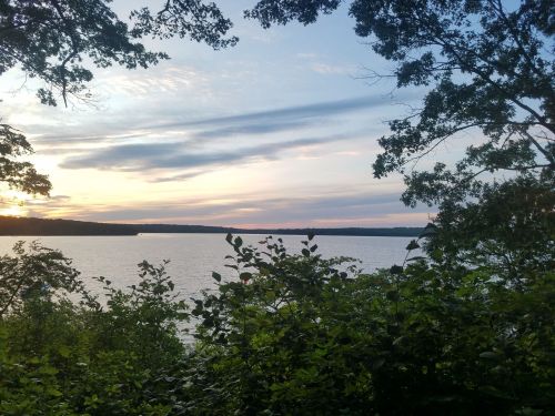 A serene lakeside view at sunset, framed by trees and foliage, with soft clouds in the sky reflecting warm colors.