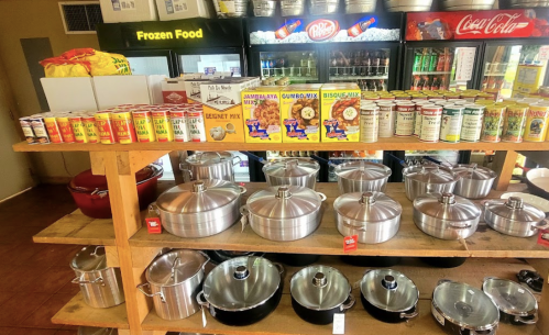 A wooden shelf displays various canned goods, cooking mixes, and metal pots in a store setting. Refrigerators in the background.