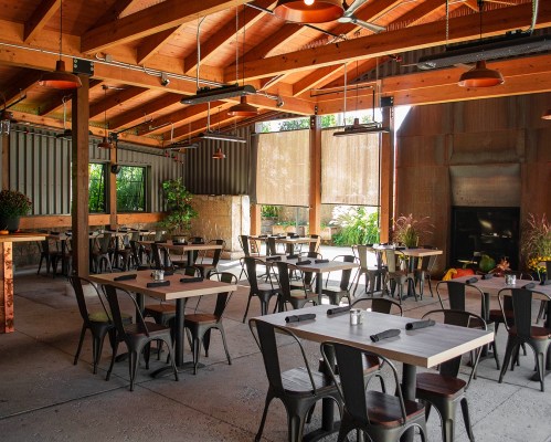 A spacious, modern restaurant interior with wooden beams, metal tables, and plants, featuring large windows and warm lighting.