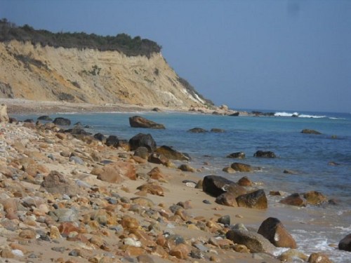 A rocky beach with smooth stones, gentle waves, and a cliff in the background under a clear blue sky.