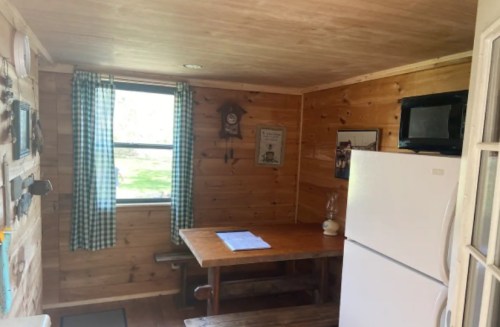 Cozy wooden cabin interior with a table, fridge, and window with checkered curtains.