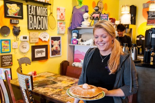 A smiling waitress holds a large pancake topped with butter in a colorful, quirky café filled with eclectic decor.
