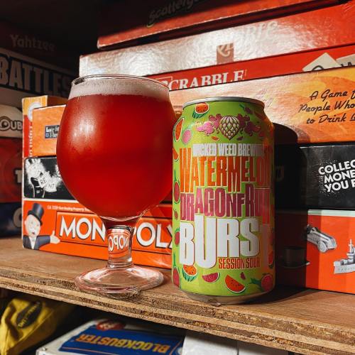 A glass of pink sour beer next to a colorful can labeled "Watermelon Dragonfruit Bursts" on a shelf of board games.