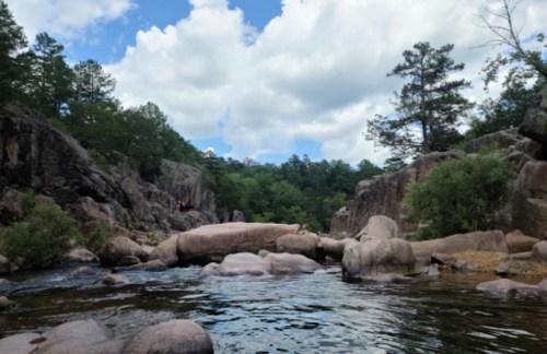 A serene river scene surrounded by rocky cliffs and lush greenery under a partly cloudy sky.