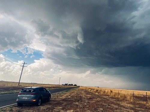 A dark, stormy sky looms over a rural road with a parked car beside a field. Power lines stretch into the distance.